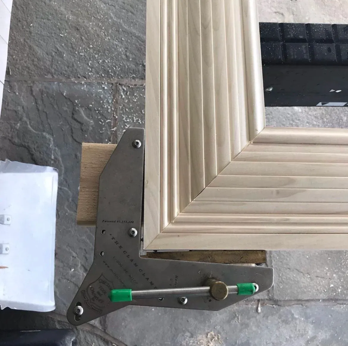 Close-up of a corner of a wooden frame being measured with a metal tool on a textured stone surface.