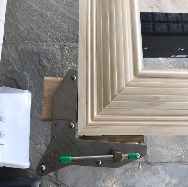 Close-up of a corner of a wooden frame being measured with a metal tool on a textured stone surface.