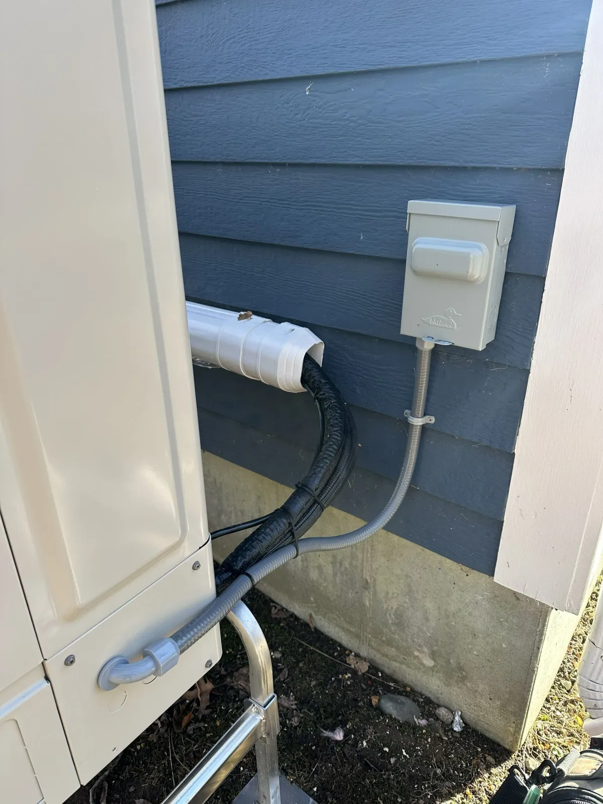 Outdoor HVAC unit connected to a gray exterior wall with a white insulated pipe and an electrical disconnect box.