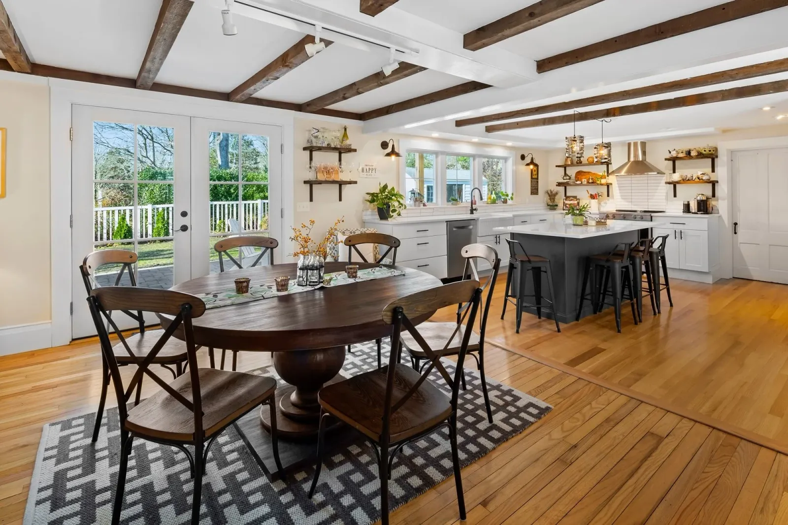 Bright modern kitchen with white cabinets, black accents, and a large island, adjacent to a dining area with a wooden table and black chairs. Sliding glass doors lead outside to a patio, and windows above the sink provide natural light. Wooden beams