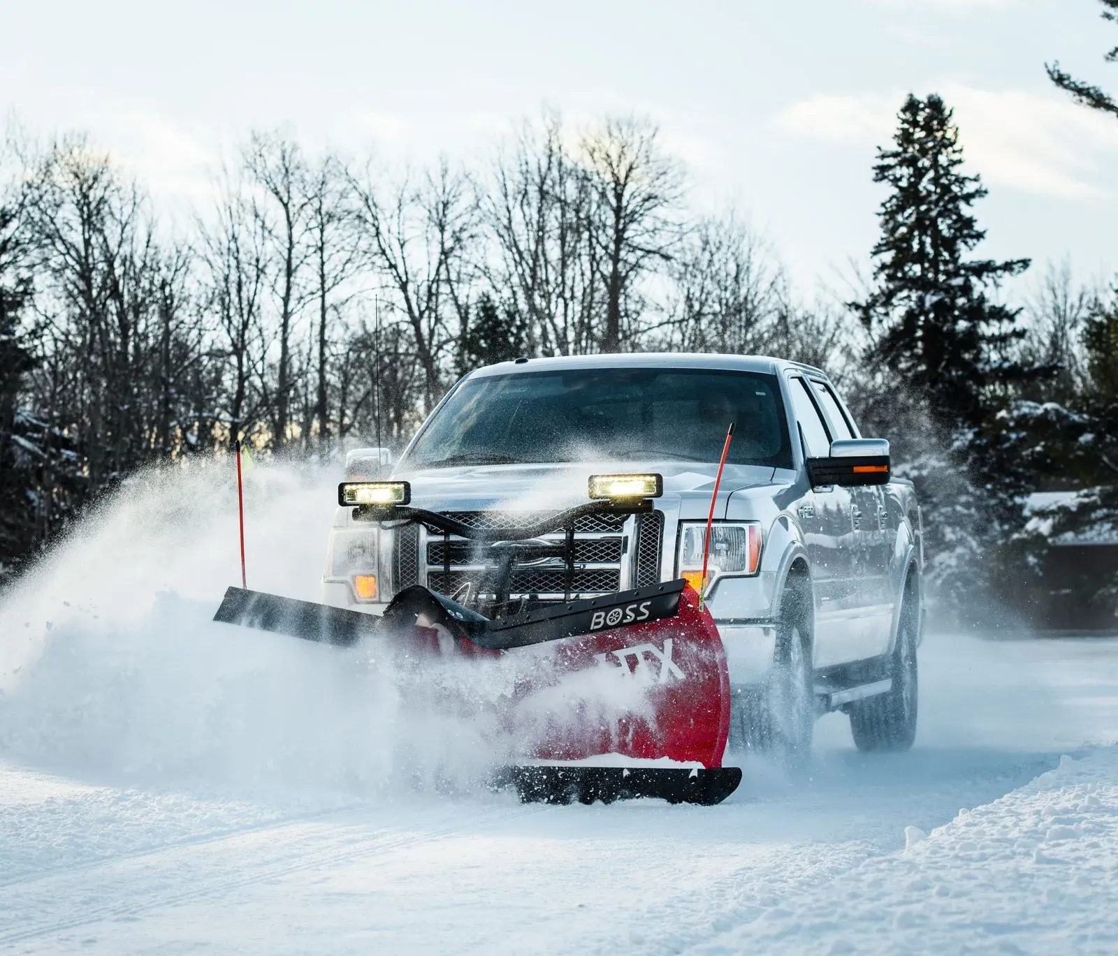 Pickup truck pushing a red snow plow through a snowy road, spraying snow