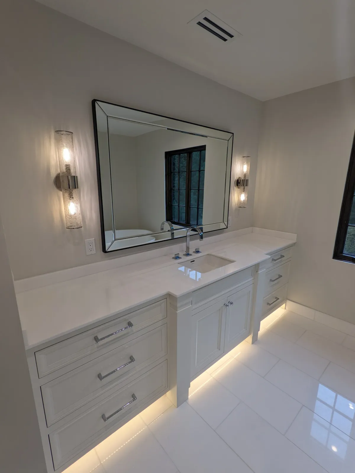 Modern bathroom vanity with white cabinetry, a large rectangular mirror with a beveled edge, two wall-mounted light fixtures, a small sink with a chrome faucet, and under-cabinet lighting on a glossy tiled floor.
