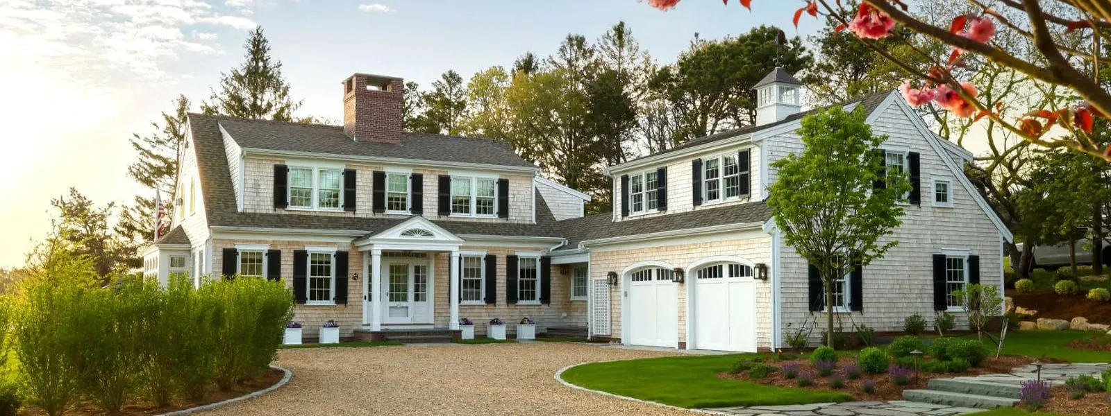 Two-story gray house with white trim and black shutters, garage, shrubs, and flowers – McPhee Associates of Cape Cod.