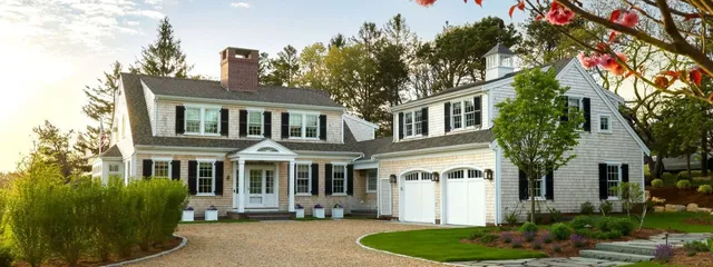 Two-story gray house with white trim and black shutters, garage, shrubs, and flowers – McPhee Associates of Cape Cod.