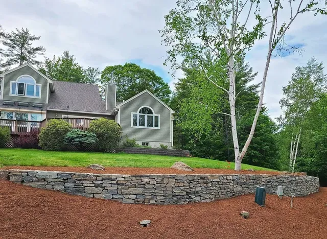 A modern home with wood siding and a arched window sits behind a stone retaining wall on a landscaped, mulched lawn.