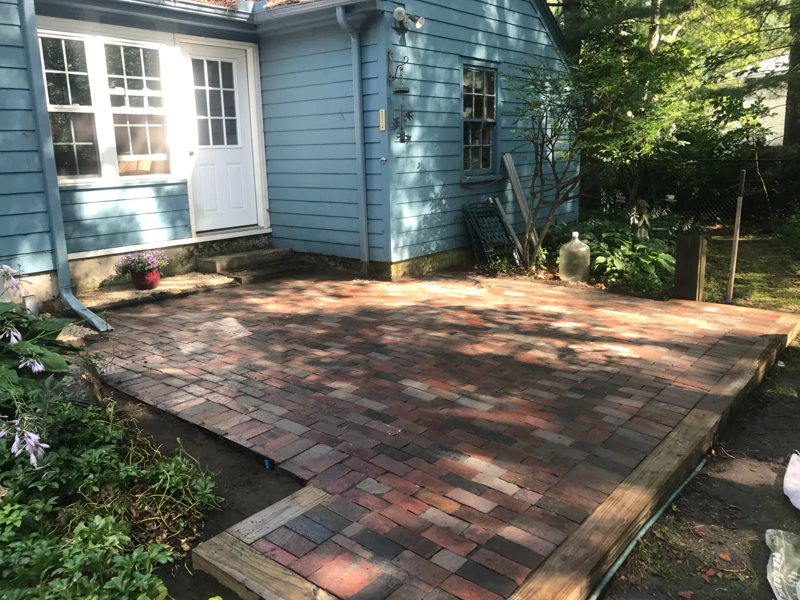 Backyard patio with red brick paving, next to a blue house, with a white door and windows, surrounded by plants and trees.