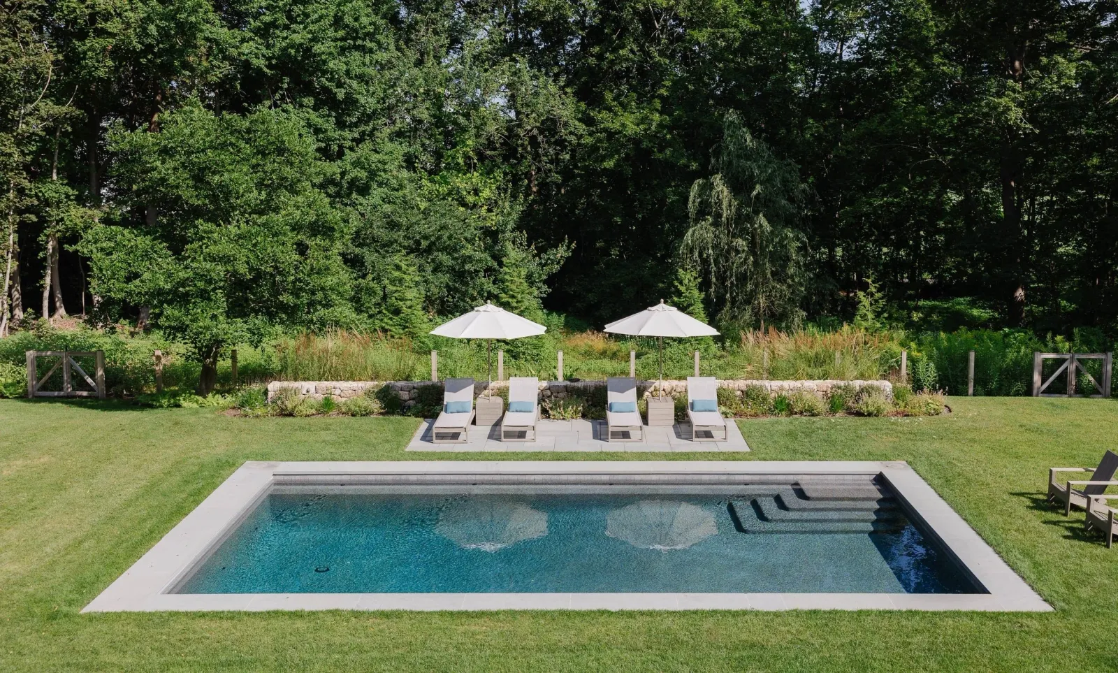 A backyard with a rectangular swimming pool, four lounge chairs with blue towels, and two white umbrellas surrounded by green grass, plants, and trees.