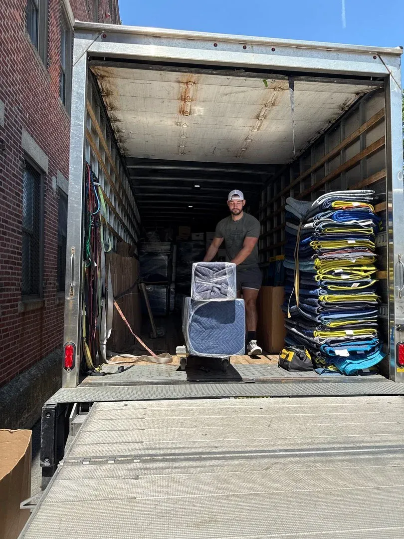 A man in a black shirt and cap unloading a trailer filled with furniture and packed items on a sunny day.