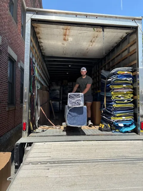 A man in a black shirt and cap unloading a trailer filled with furniture and packed items on a sunny day.