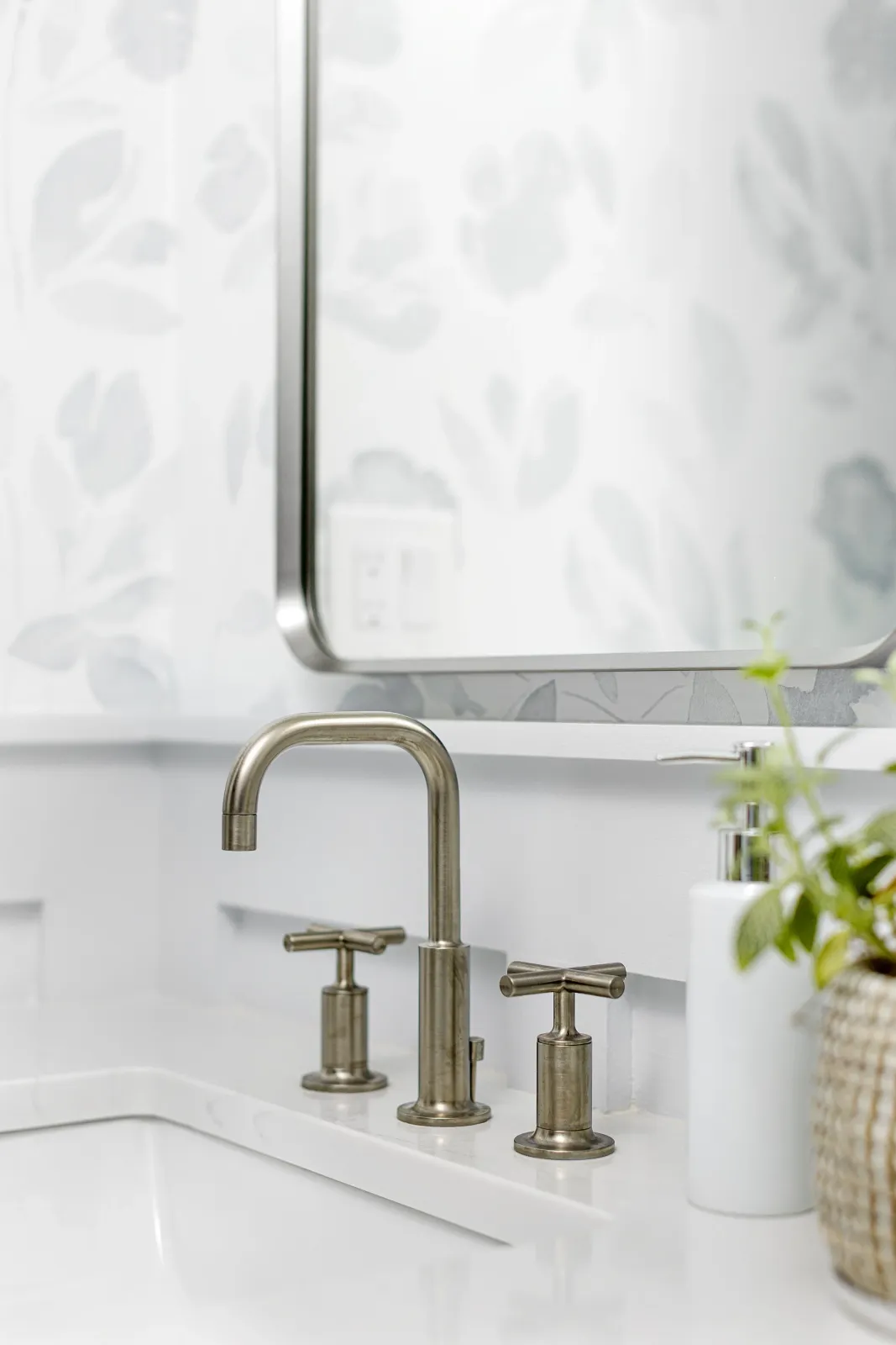 Close-up of a modern bathroom sink with a brushed metal faucet and handles, white countertop, a soap dispenser, and a potted plant, with a mirror and a floral patterned wall in the background.