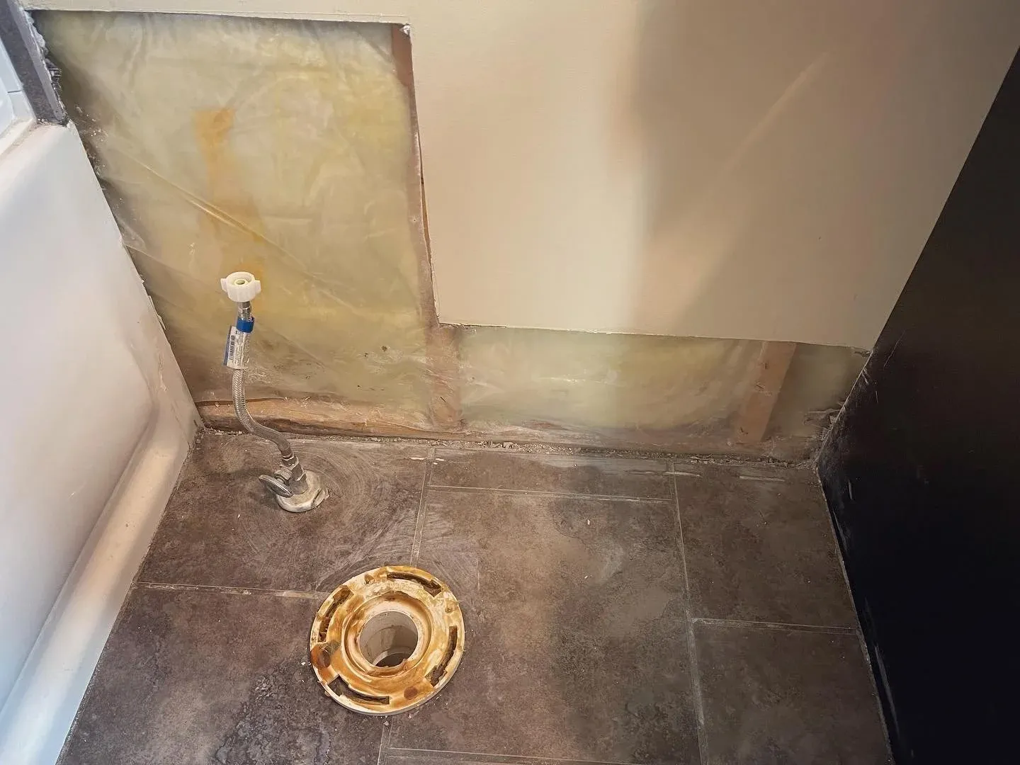 Bathroom floor with exposed plumbing and a removed toilet, showing a drain flange on the floor and wall insulation behind the partially removed wall.