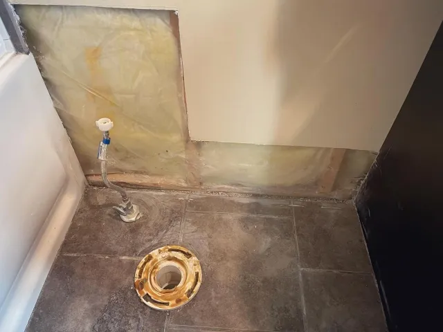 Bathroom floor with exposed plumbing and a removed toilet, showing a drain flange on the floor and wall insulation behind the partially removed wall.