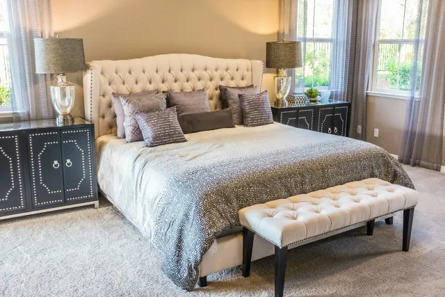 A bedroom with a large bed featuring an upholstered, tufted headboard, adorned with multiple decorative pillows in shades of gray. There are two black nightstands with white decorative patterns on either side of the bed, each holding a large glass lamp with a gray lampshade. The room has a beige wall, large windows with sheer curtains, and a beige carpet.