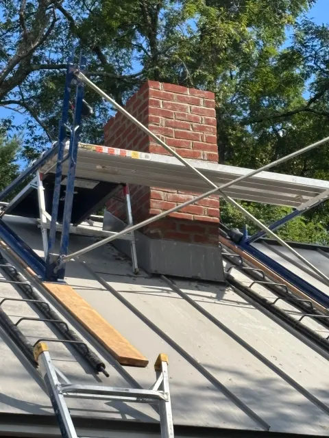 A rooftop with a brick chimney, scaffolding, and ladders for roof work, surrounded by trees and a blue sky.