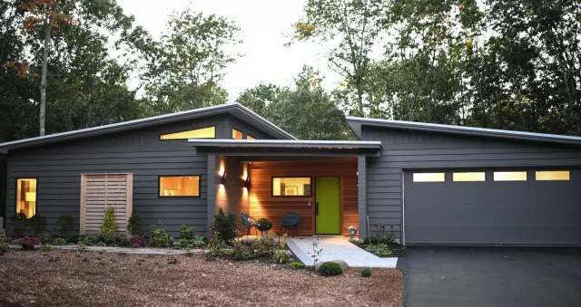 Modern single-story gray house with a sleek design, featuring wooden accents, large windows, and a double garage. Surrounded by trees and landscaped yard.