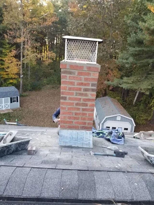 Brick chimney on a shingled roof with a metal cap, surrounded by trees in the background.