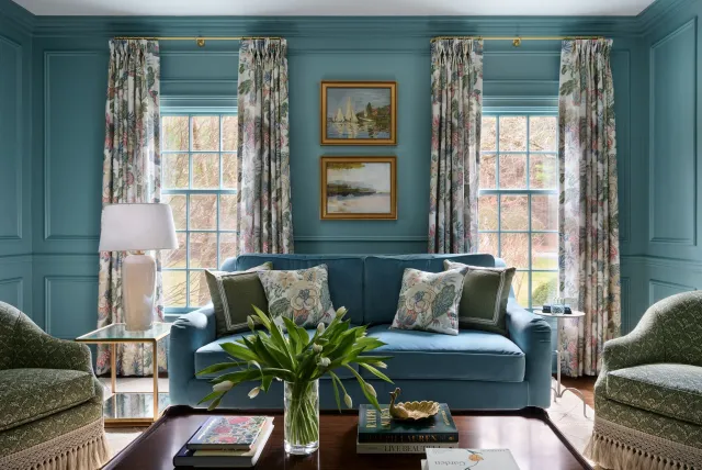 Living room with blue walls, curtains with floral pattern, blue sofa with floral and green pillows, two armchairs with green upholstery, a glass vase with white tulips on a wooden coffee table, paintings on the wall, and view of trees outside the win