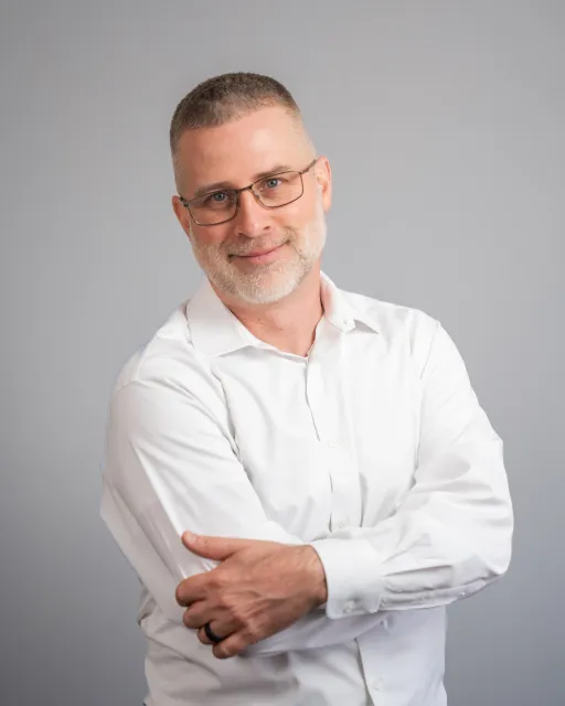 A middle-aged man with glasses, short hair, and a beard, wearing a white dress shirt, smiling, standing with arms crossed against a plain gray background.