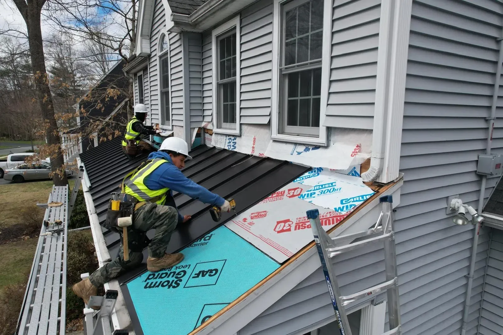 Workers install roofing underlayment on a house with gray siding and dormer windows.