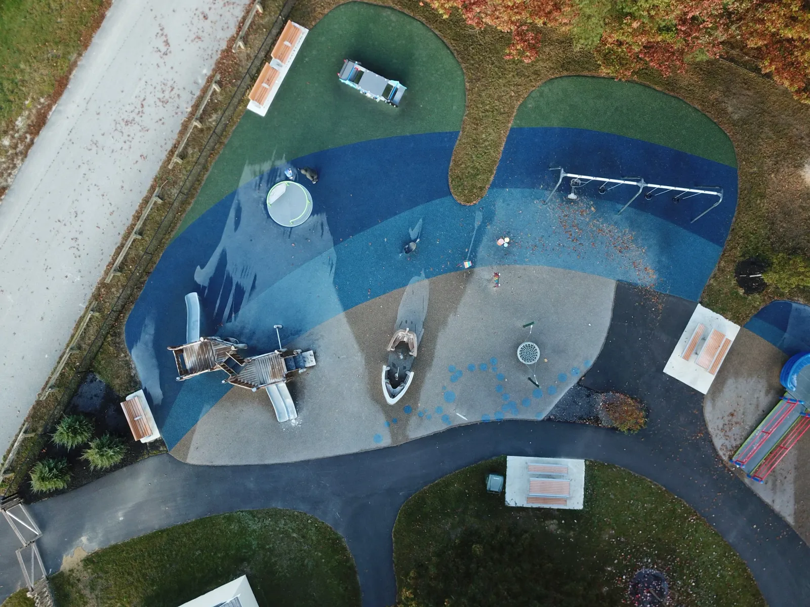 A bird's eye view of a colorful playground with swings, slides, a trampoline, and benches, surrounded by a paved pathway and trees.