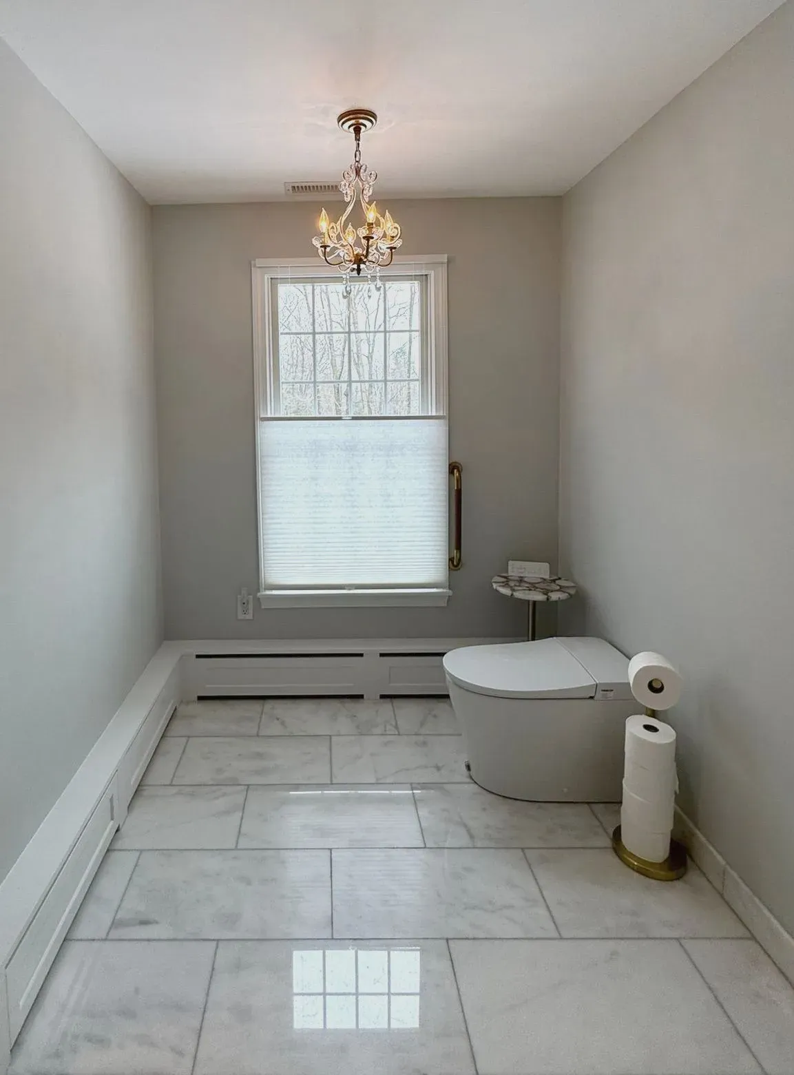 Empty modern bathroom with white tile floor, chandelier, window, and toilet paper holder.