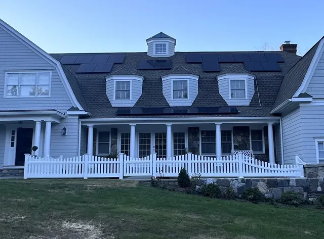 White vinyl scalloped picket fence in front of a large shingled colonial home with stone retaining wall and covered porch.
