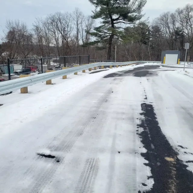 Snow-covered road with metal guardrail. Trees in the background, a small building to the right.