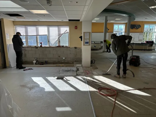 Renovation work in a bright room with two workers, tools, exposed floor, and debris near windows.