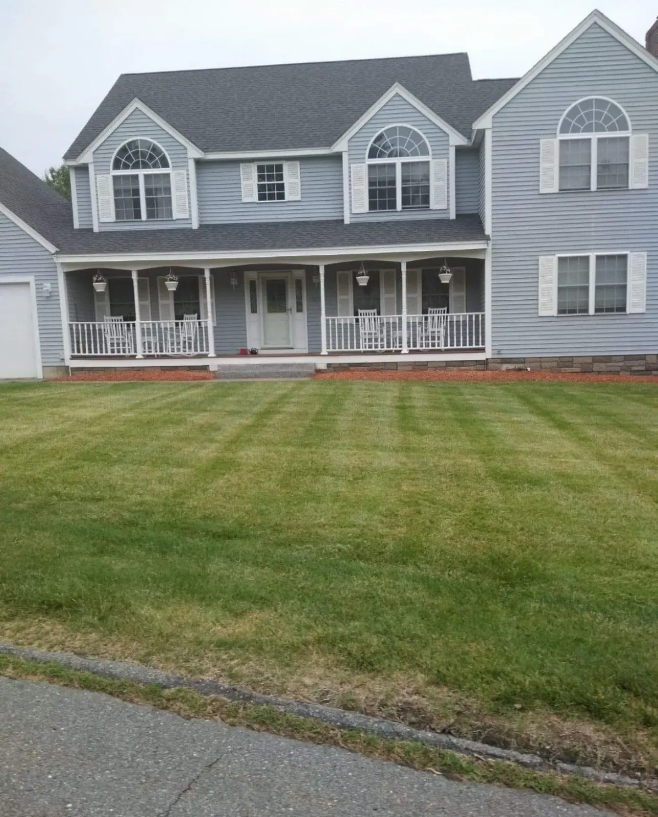A light gray, two-story house with a front porch, a gabled roof, and a mowed lawn under an overcast sky.