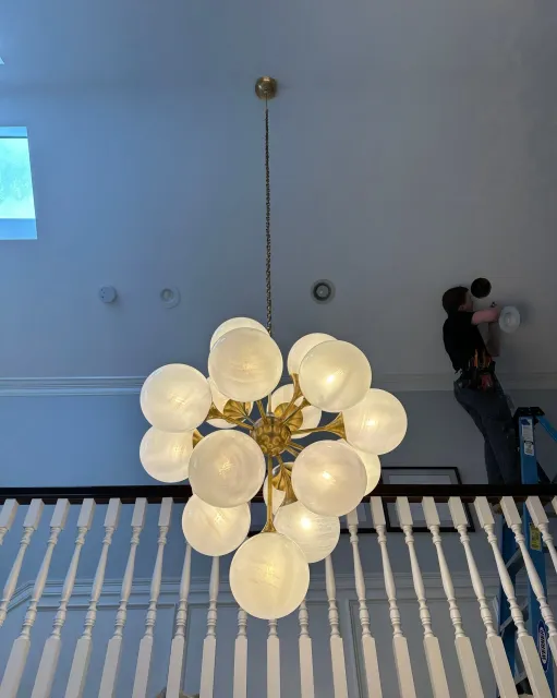 A chandelier with multiple glowing white globes hanging from a ceiling. A person is on a ladder, working near the ceiling.