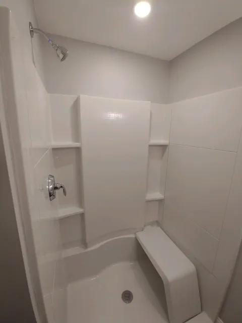 Empty shower stall with white tiles, chrome showerhead, faucet, and small built-in shelves, with a curved white seat.