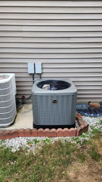 An outdoor air conditioning unit installed beside a house with beige siding. The unit is gray and black, with a fan on top, sitting on a concrete pad bordered by small red bricks and white pebbles. Adjacent is a white unit and a small piece of brick