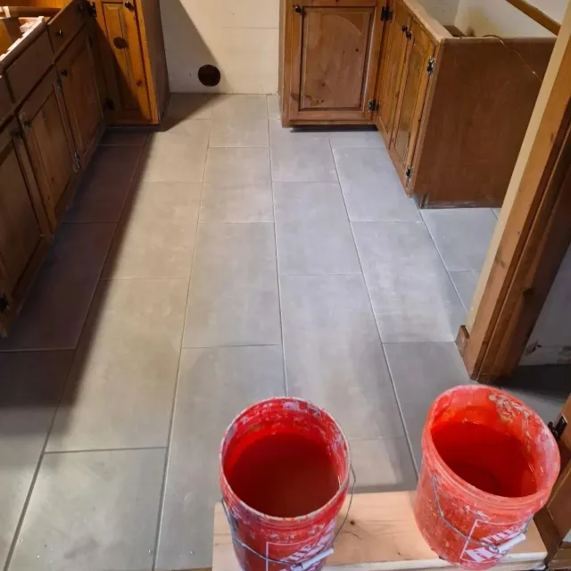 Interior of a kitchen under renovation with beige tiled floor, wooden cabinets, and two buckets the foreground.