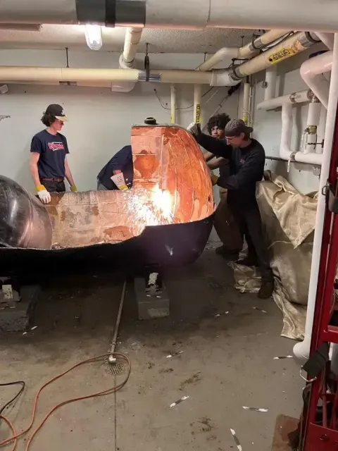 Four people in a basement workshop work on a large, partially cut-open dark-colored metal boiler or tank.