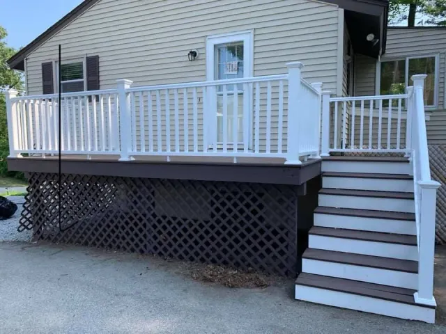 A raised deck with white railings and stairs, a brown wooden base, and brown-and-white steps, attached to a beige house.