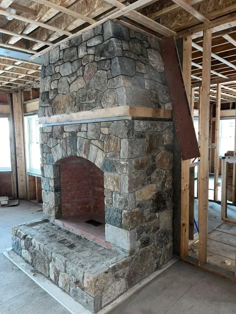 Construction site with a stone fireplace under construction, surrounded by exposed wooden framing and unfinished interior walls.