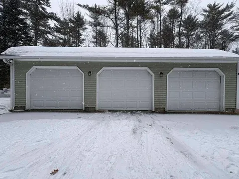 A three-car residential garage with white doors attached to a light-colored house, surrounded by evergreen trees with snow on the ground