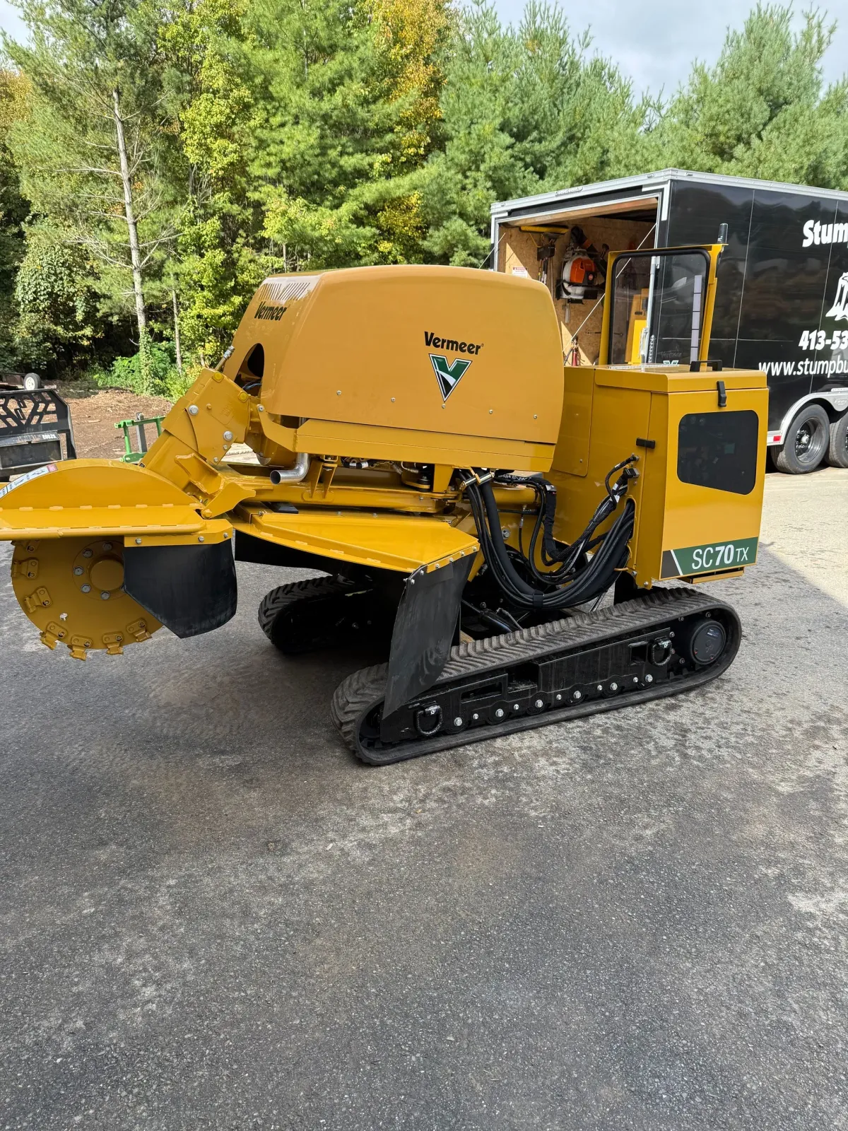 Yellow and black stump grinder on tracks, parked on pavement, next to a trailer.