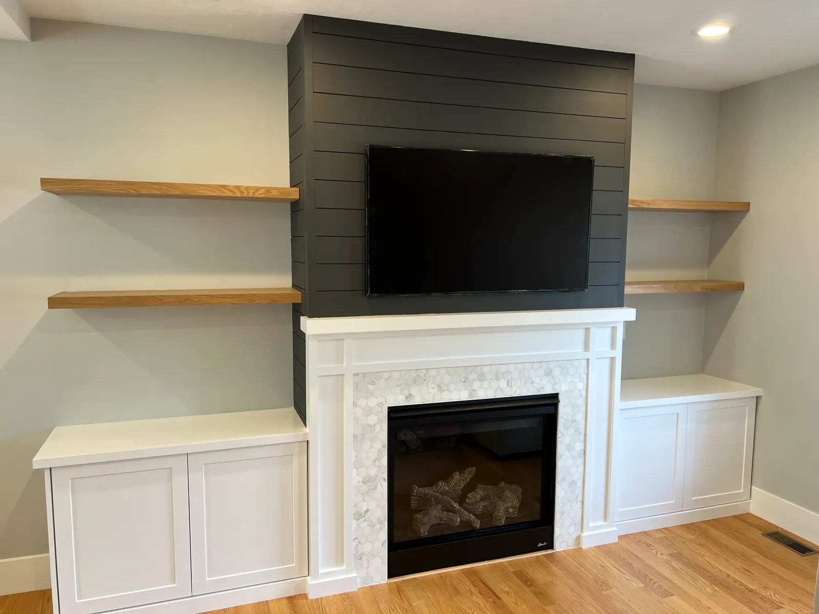 Modern living room featuring a sleek, black accent wall with a mounted television, flanked by wooden floating shelves. Below, a contemporary white fireplace with a stone facade and cabinetry on either side.