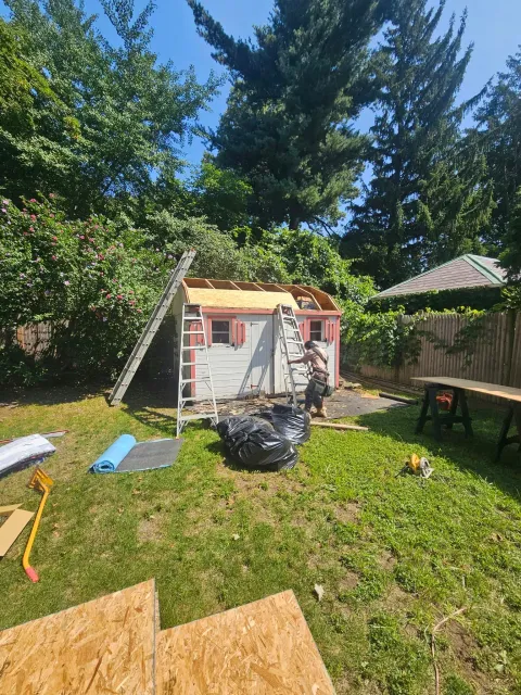 Backyard playhouse under tall trees, with lawn tools and a table nearby.