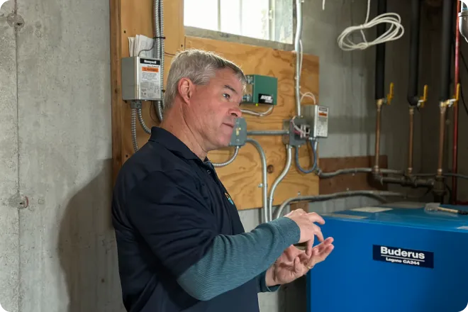 A man stands in a utility room near electrical panels and a blue Buderus boiler, gesturing with his hands while speaking.