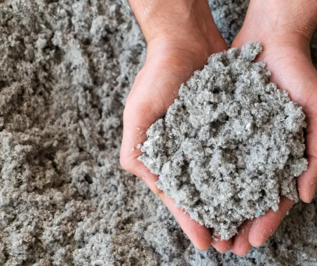 Hands holding a large clump of Cellulose insulation, recycled paper and treated for fire and pest resistance. It’s dense, effective at sealing air leaks, and ideal for attics and walls.
