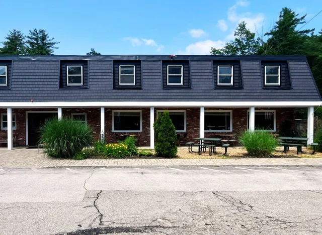 Two-story brick building with black siding and outdoor picnic tables among greenery