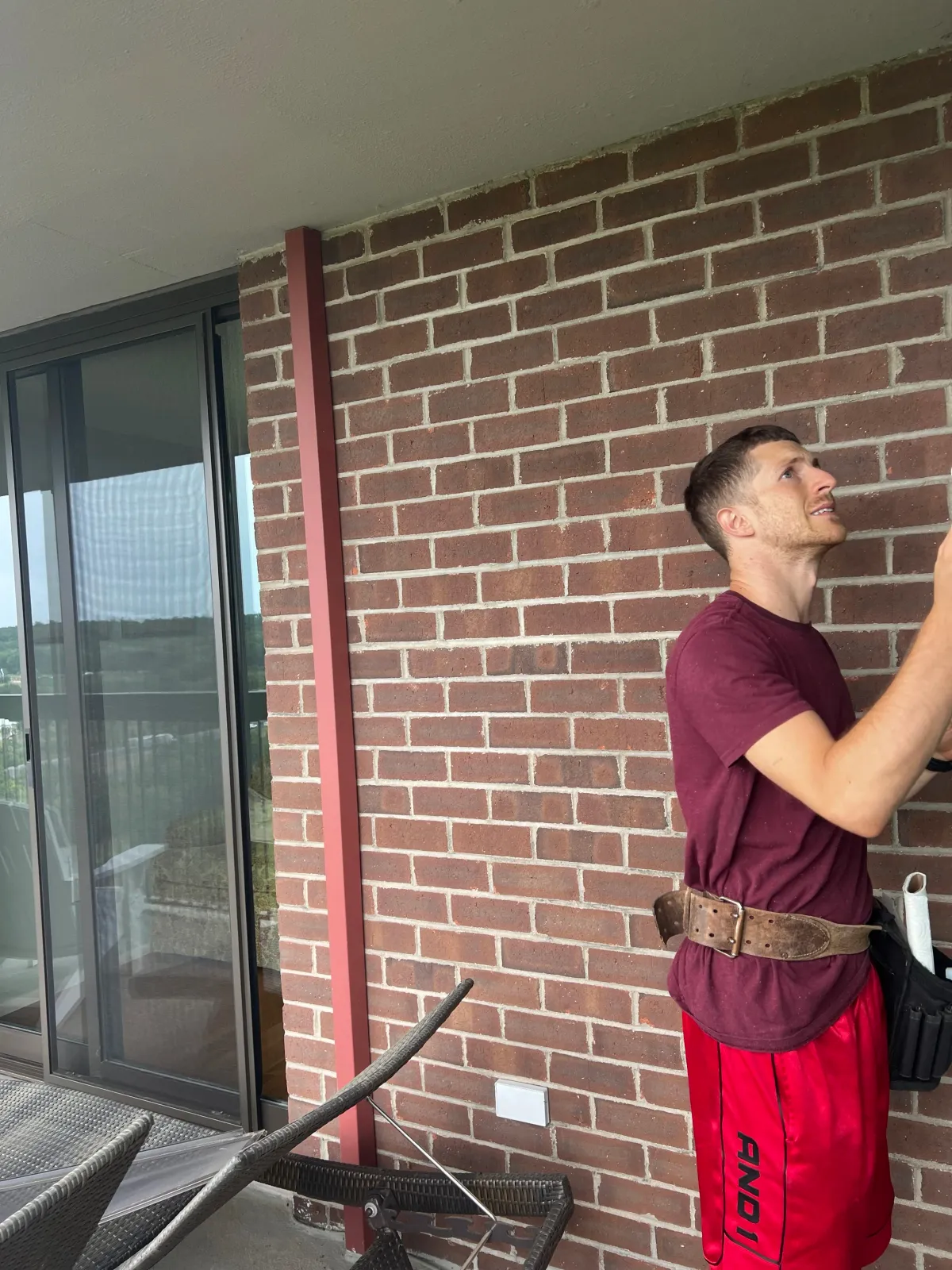 Person standing on a balcony with a brick wall background, wearing a maroon t-shirt and red shorts, holding a tool with a belt and pouch around the waist, next to a sliding glass door and outdoor furniture.