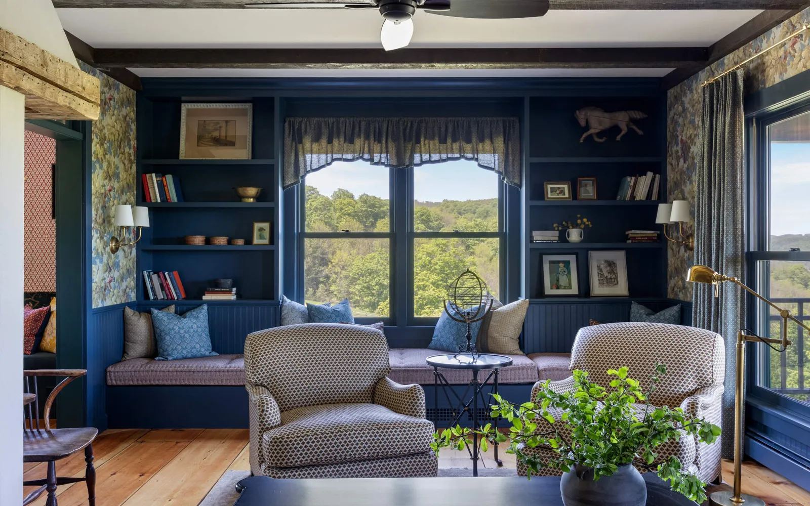 Wide view of the living area showcasing a deep red sofa with a mustard throw blanket and patterned pillows. The room features dark wood ceiling beams, extensive floral wallpaper above wainscoting, and a staircase leading to an upper level visible in the background.