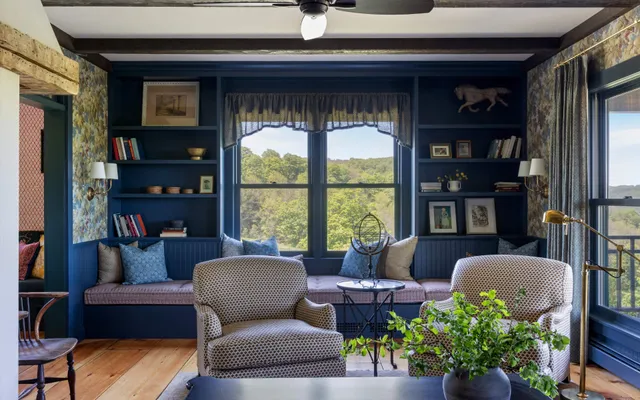 Wide view of the living area showcasing a deep red sofa with a mustard throw blanket and patterned pillows. The room features dark wood ceiling beams, extensive floral wallpaper above wainscoting, and a staircase leading to an upper level visible in the background.