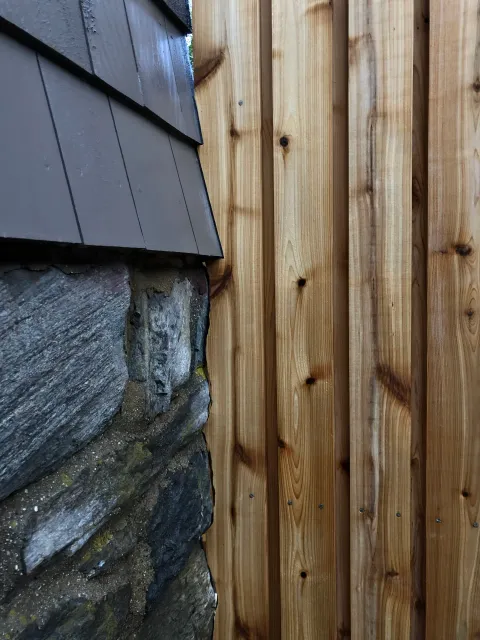 Close-up view of an exterior corner of a building showing a stone wall, wooden fence, and part of the siding.