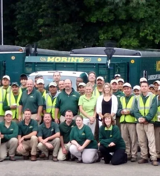 A large group of people in green safety vests stands in front of multiple trucks, with trees in the background.