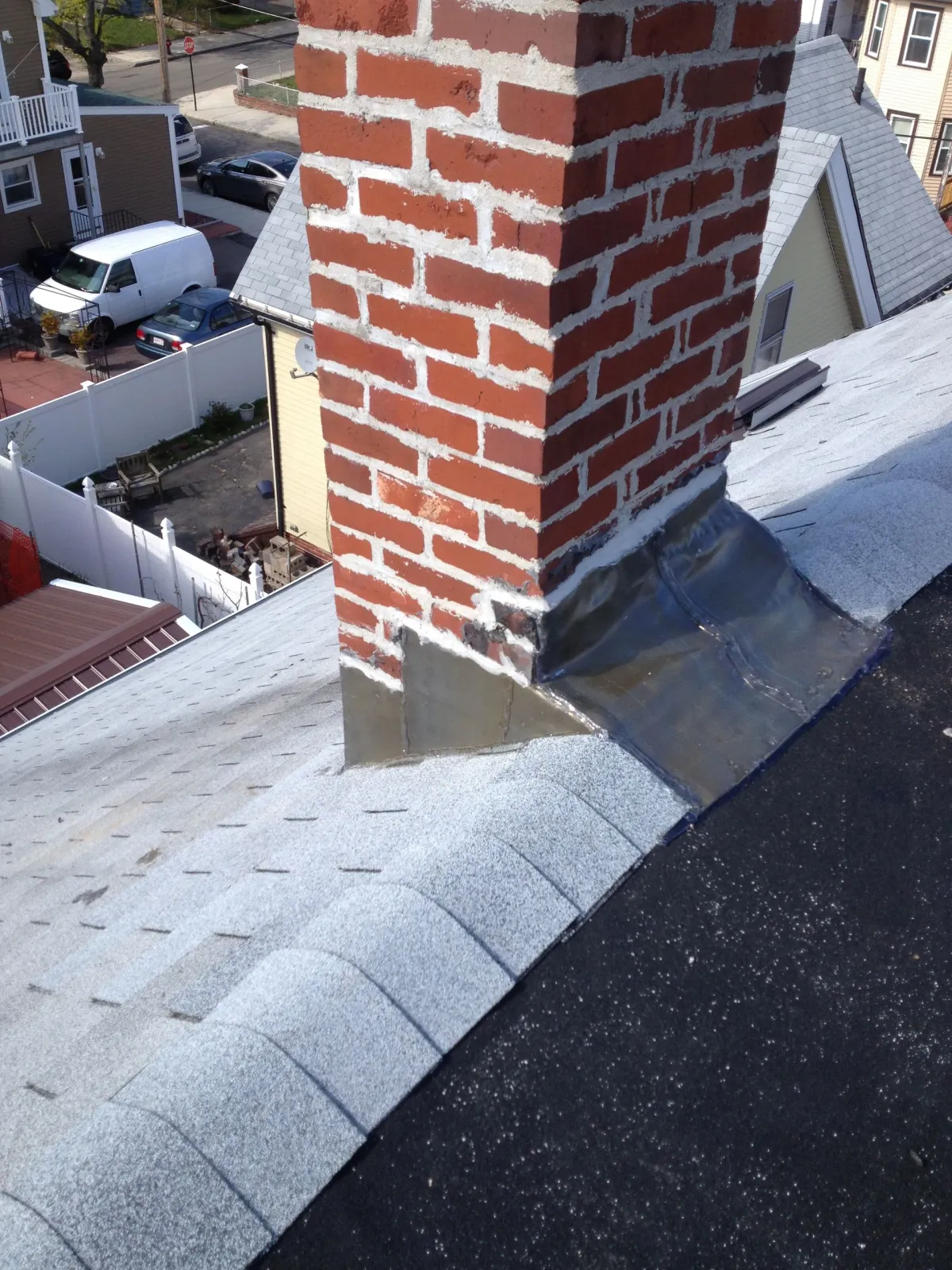 Red brick chimney rising through a gray shingled rooftop with nearby houses visible below