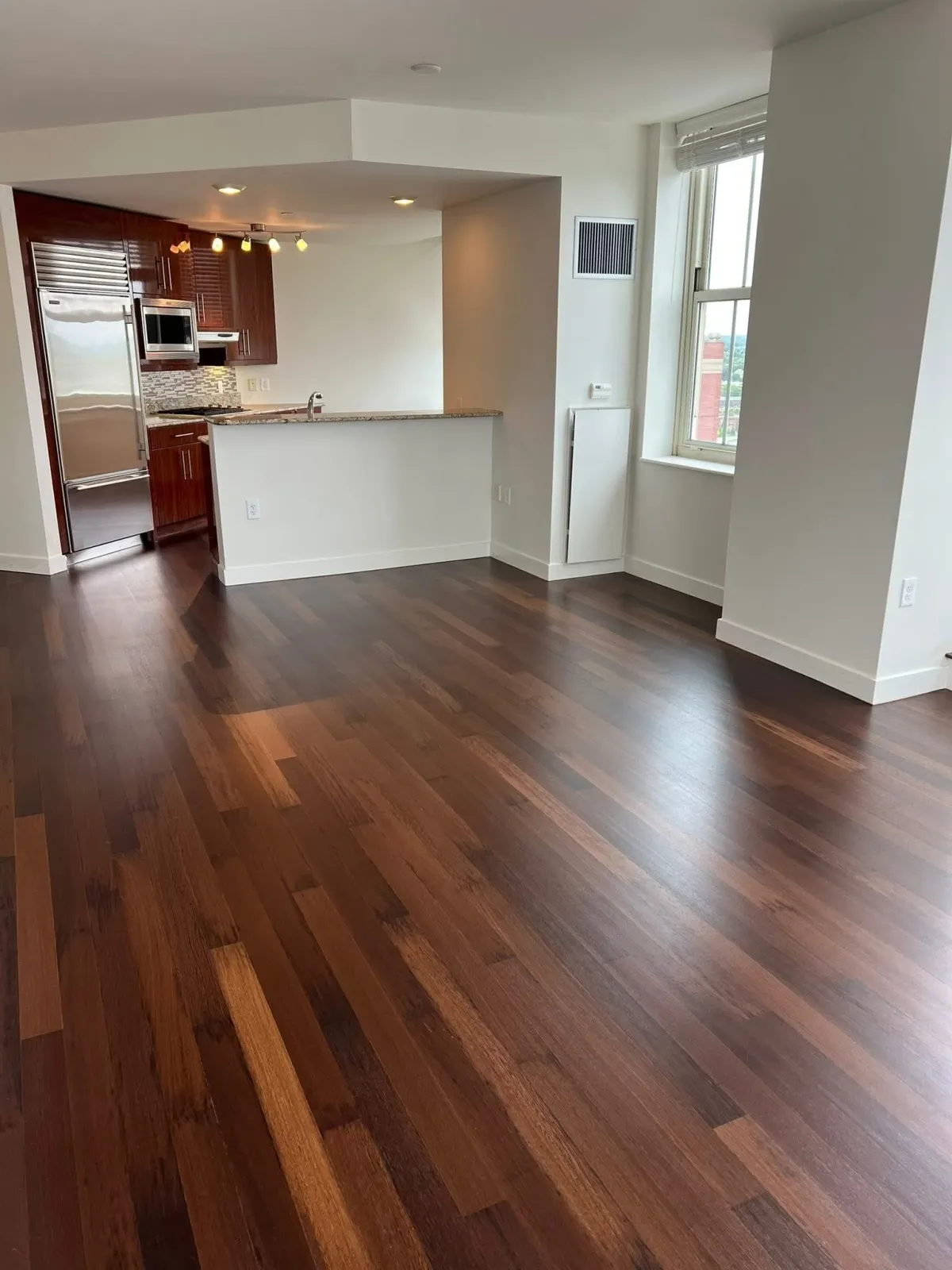 Empty living space with dark hardwood floors, white walls, and large windows letting in natural light. The partially visible kitchen has reddish-brown cabinets, a stainless steel refrigerator, and a granite countertop.